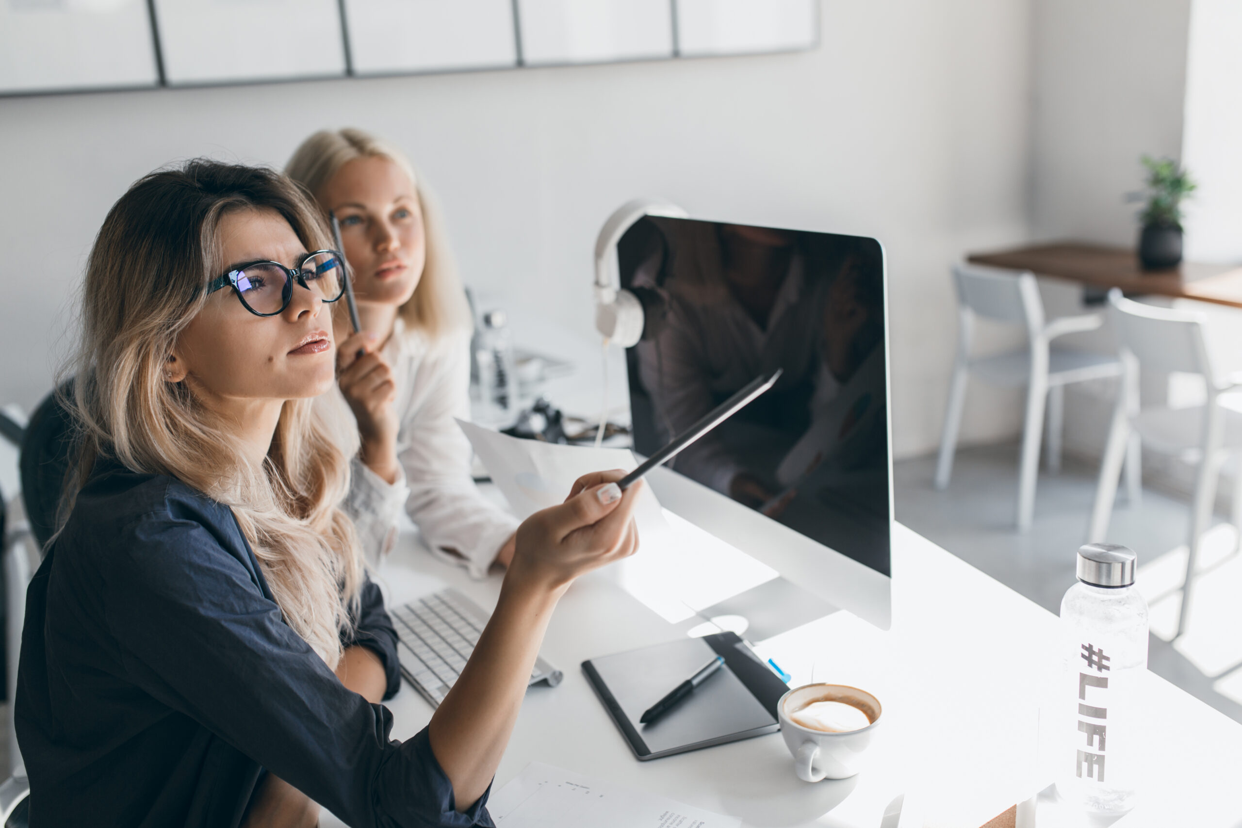 Thoughtful blonde woman in glasses holding pencil and looking away during work in office. Indoor portrait of busy long-haired female accountant using computer..